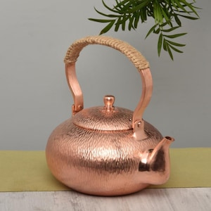 May include: A copper teapot with a textured surface and a woven handle. The teapot has a rounded body, a spout, and a lid with a small knob. The handle is made of copper and wrapped with a natural fiber. The teapot is a warm copper color.