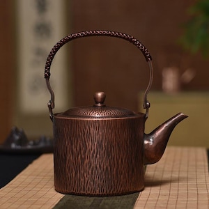 Może przedstawiać: Miedziany czajnik z teksturowaną powierzchnią i plecionym uchwytem. Czajnik posiada dzióbek i pokrywkę z małym pokrętłem. Uchwyt jest przymocowany do korpusu czajnika za pomocą metalowych wsporników. Czajnik stoi na bambusowej macie.