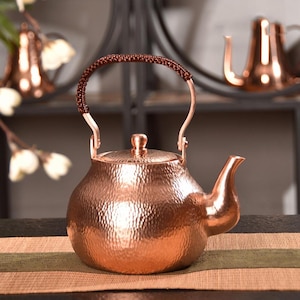 May include: A copper-colored teapot with a hammered texture. The teapot has a curved spout, a lid with a small knob, and a handle wrapped in a brown, braided material. The teapot is sitting on a brown and green striped surface.