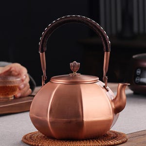 Puede incluir: Una tetera de color cobre con un asa marrón oscuro y una tapa decorativa. La tetera tiene un cuerpo redondeado con un diseño facetado y un pico curvo. El asa está envuelta con un material de color más claro.