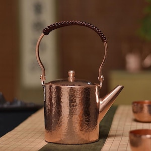 May include: A copper-colored teapot with a hammered texture. It has a curved handle with a woven brown grip and a small, pointed spout. The lid is topped with a small knob. The teapot is designed for tea service.