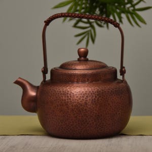 May include: A copper-colored teapot with a hammered texture. The teapot has a curved spout, a lid with a small knob, and a handle with a woven grip. The teapot is designed for brewing tea.