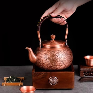 Puede incluir: Una tetera de cobre con textura martillada, asa marrón oscuro y tapa a juego. La tetera se asienta sobre una base de madera con un mando. Una pequeña bandeja de madera contiene hojas de té y una taza de cobre está cerca.