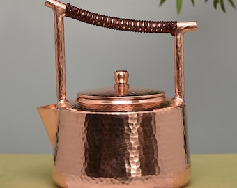 May include: A copper tea kettle with a hammered texture. The kettle has a spout, a lid with a small knob, and a handle wrapped in a brown, woven material. The kettle is approximately 10 inches tall.