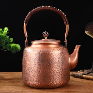 May include: A copper-colored teapot with a hammered texture. It has a curved handle wrapped in brown material and a small decorative knob on the lid. The spout is curved, and the teapot sits on a wooden surface.