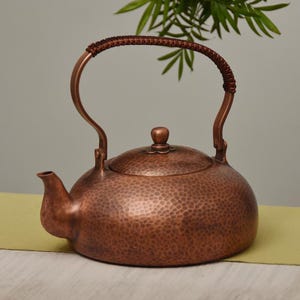 May include: A copper teapot with a hammered texture and a woven handle. The teapot has a curved spout and a lid with a small knob. The handle is made of copper and woven material. The teapot is a warm, reddish-brown colour.