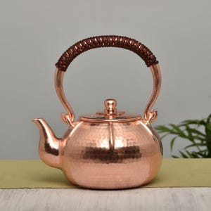 Può includere: Una teiera in rame con una texture martellata e un manico intrecciato marrone. La teiera ha un beccuccio curvo e un coperchio con un piccolo pomello. La teiera è progettata per preparare il tè.
