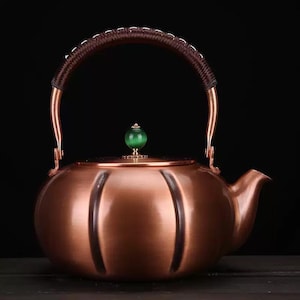 May include: Copper teapot with a brown woven handle and a green gemstone knob. The teapot has a rounded body with vertical grooves and a spout for pouring. The handle is arched, and the teapot sits on a dark surface.