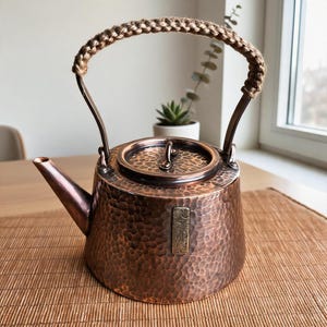 May include: A hammered copper teapot with a rounded body, a short spout, and a lid featuring a small handle. The arched metal handle is wrapped in a thick, braided tan jute rope. A rectangular brass plaque with engraved Asian characters is attached.