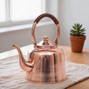 May include: A polished copper tea kettle with a rounded body, a curved spout, and a lid with a small knob. The handle is arched and wrapped in a dark brown textured material, and the lower section of the kettle features horizontal line detailing.