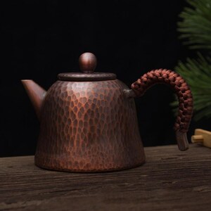 May include: A copper-coloured teapot with a textured surface and a woven handle. The teapot has a small spout and a rounded lid with a small knob. The handle is a dark brown colour. The teapot is on a wooden surface.