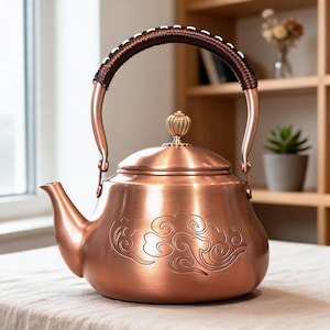 Pode incluir: Uma chaleira de metal cor de cobre com um corpo arredondado e um padrão de nuvens gravado. A chaleira tem um bico curvo, uma tampa removível com um pequeno botão dourado e uma alça alta arqueada envolta em material de corda castanho.
