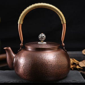 Puede incluir: Una tetera de color cobre con una textura martillada. Cuenta con un asa dorada y un pomo decorativo plateado en la tapa. El pico está en el lado izquierdo. La tetera está diseñada para preparar té.