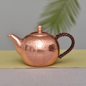 May include: A copper teapot with a hammered texture. The teapot has a curved spout, a lid with a small knob, and a braided brown handle. The teapot is sitting on a yellow surface.