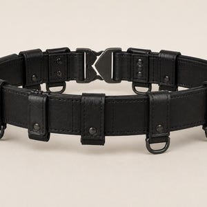May include: A black utility belt featuring a matte finish, multiple vertical loops, and several small metal D-rings attached along the lower edge. The belt is secured with a central black metal buckle mechanism that interlocks at the front.