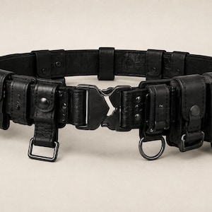 May include: A black utility belt constructed from textured leather, featuring multiple small pouches with snap-button closures, integrated metal D-rings, and a central matte black tactical-style quick-release buckle mechanism.