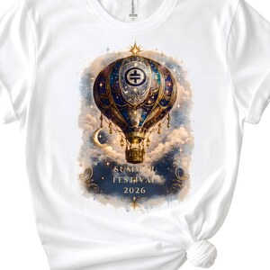 May include: A white t-shirt featuring a graphic of a blue and gold hot air balloon floating among clouds, stars, and a crescent moon. The balloon has a circular emblem with a symbol. Below the balloon, the text reads SUMMER FESTIVAL 2026.