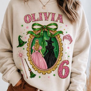 Op de afbeelding: Een beige sweatshirt met de naam OLIVIA in roze glittertekst boven een gouden ovaal frame met twee figuren in roze en zwarte jurken. Het ontwerp bevat een groene strik, een heksenhoed, een toverstaf, een bezem, een tiara en het cijfer 6.