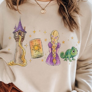 May include: A beige crewneck sweatshirt featuring a graphic print of a tall stone tower with a purple roof, a long golden braid, a glowing lantern with a sun emblem, a person with long blonde hair in a purple dress, and a green chameleon.