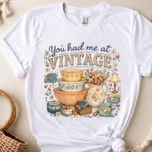 May include: White t-shirt featuring a vintage-themed graphic. The design includes the text "You had me at VINTAGE" above a collection of vintage kitchenware, a floral pitcher, and retro telephones. The design is in shades of blue, orange, and yellow.