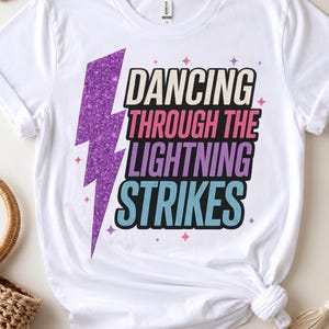Pode incluir: Uma t-shirt branca de manga curta com um raio roxo brilhante no lado esquerdo e o texto DANCING THROUGH THE LIGHTNING STRIKES em linhas sobrepostas nas cores creme, rosa, roxo e azul claro com pequenos detalhes de estrelas.