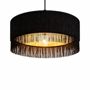 Può includere: Un lampadario nero e rotondo con un paralume a frange. Il paralume ha un aspetto strutturato e circonda un interno caldo e dorato. La luce è appesa a un cavo nero, creando un elemento di illuminazione decorativo moderno.