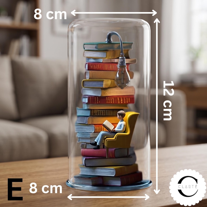 May include: A glass cloche containing a stack of colorful books, a miniature figure sitting in a yellow armchair reading, and a small lamp. The display measures 8 cm in width and 12 cm in height. The text CLART&Eacute; is visible in the bottom right corner.