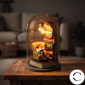 May include: A decorative glass cloche containing a miniature scene of a person sitting in a yellow armchair reading a book. A stack of books stands beside the chair, illuminated by a small hanging light bulb. The entire display rests on a large book.