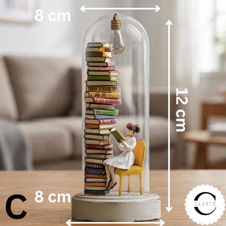 May include: A decorative glass cloche containing a tall stack of multicolored books and a figurine sitting on a yellow chair reading a book. A lightbulb hangs from the top inside. The base is white, and the dimensions are 8 cm by 12 cm.