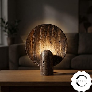 May include: A circular table lamp crafted from brown travertine stone with a natural porous texture. The lamp features a large, flat disc-shaped shade mounted on a cylindrical base, emitting a warm, soft glow from behind the disc.