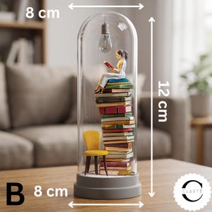 May include: A glass cloche display containing a stack of multicolored books, a yellow chair, and a figurine reading a book under a hanging light bulb. The base is gray. Dimensions are 8 cm in width and 12 cm in height. The letter B is visible.