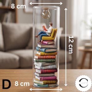 May include: A cylindrical glass display containing a stack of colorful books with a figure sitting on a yellow chair reading a red book. A small light bulb hangs from the top. The object measures 8 cm in width and 12 cm in height.