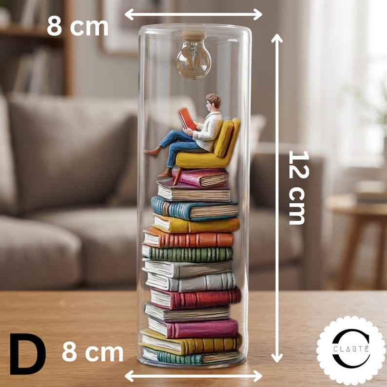 May include: A cylindrical glass display containing a stack of colorful books with a figure sitting on a yellow chair reading a red book. A small light bulb hangs from the top. The object measures 8 cm in width and 12 cm in height.