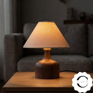 May include: A table lamp featuring a dark brown wooden base with a smooth, rounded shape and a brass-colored neck. The lamp is topped with a conical, textured beige fabric shade that emits a warm, soft glow. A logo with the text CLARTÉ is visible.
