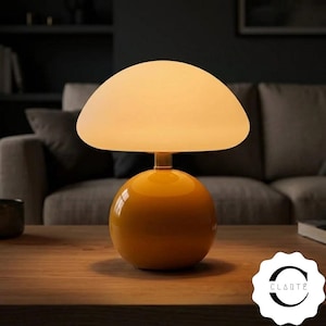 May include: A table lamp with a spherical, glossy mustard yellow base and a wide, mushroom-shaped frosted white glass shade that emits a warm glow. The base connects to the shade with a small metallic neck. A logo with the text CLARTÉ is visible.