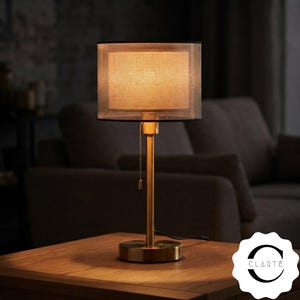 May include: A table lamp with a cylindrical, textured beige fabric shade and a metallic gold-toned base. The lamp features a thin vertical stem and a pull chain switch. The light emits a warm glow, illuminating the shade and the surface below.