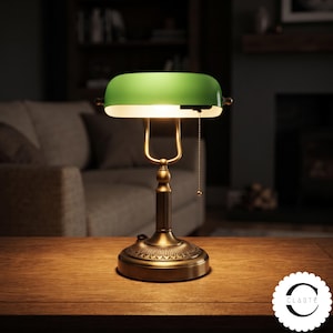 Puede incluir: Una lámpara de escritorio con una pantalla de cristal verde brillante y una base de metal en tono latón con una columna estriada y un pedestal circular decorativo. La lámpara incluye un interruptor de cadena y emite luz cálida bajo la pantalla.