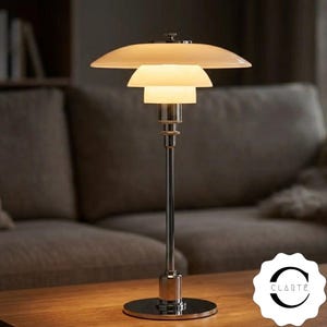 May include: A table lamp with a polished chrome metal base and stem, featuring three stacked, tiered white glass shades that diffuse light. The lamp has a circular base and a logo in the corner with the text CLARTÉ in black capital letters.