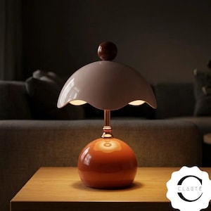May include: A table lamp featuring a glossy orange spherical base, a metallic stem, and a wide, scalloped, off-white shade topped with a dark brown sphere. The lamp emits a warm glow from beneath the shade. The text CLARTÉ is visible in the corner.