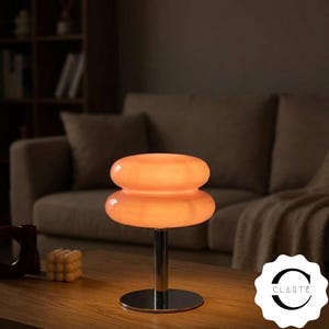 May include: A table lamp featuring a double-tiered, glowing orange glass shade with a smooth, rounded silhouette. The lamp is supported by a polished chrome cylindrical stem and a circular base. The text CLARTÉ is visible in the bottom right corner.
