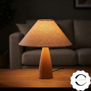 May include: A table lamp with a conical wooden base and a textured, light beige fabric lampshade that emits a warm glow. The lamp features a black power cord extending from the base. A white circular logo with the text CLARTÉ is visible.