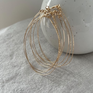 Peut inclure: Un ensemble de boucles d'oreilles créoles dorées. Les boucles d'oreilles sont fines et ont une surface texturée. Les créoles sont disposées en couches, avec plusieurs créoles imbriquées. Les boucles d'oreilles sont présentées sur une surface gris clair.