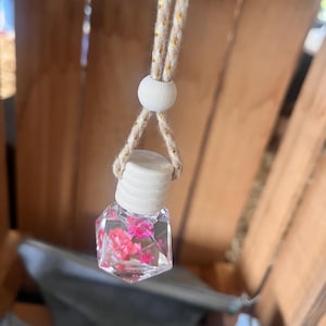 May include: A small, clear, geometric-shaped glass bottle filled with pink flowers, suspended by a natural rope cord with gold accents. The bottle has a wooden cap and a wooden bead. The item is likely an air freshener or decorative item.