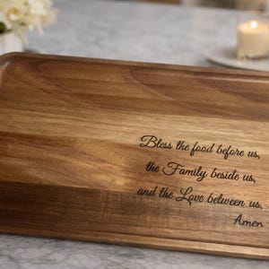 May include: A rectangular wooden serving tray with rounded corners. The tray is made of light and dark brown wood. Engraved on the tray is the text: "Bless the food before us, the Family beside us, and the Love between us, Amen."