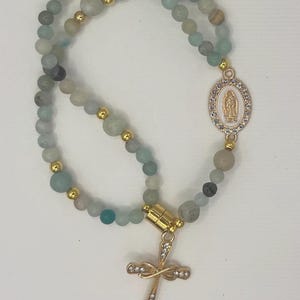 May include: A rosary necklace with light blue and gray gemstone beads. The necklace features gold-colored accents, a cross pendant with clear crystals, and a gold-colored oval pendant with a religious image.