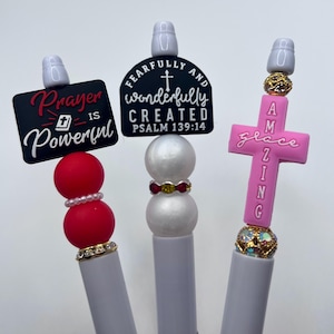 May include: Three white pens with decorative toppers. The first has a black sign reading Prayer is Powerful above red beads. The second has a black sign reading Fearfully and Wonderfully Created Psalm 139:14 above white beads. The third has a pink cross reading Amazing Grace.