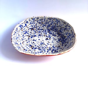 Może przedstawiać: Owalna ceramiczna miska do serwowania z białym wnętrzem, nakrapiana niebieskimi i brązowymi kropkami. Obręcz jest w żywym czerwonym kolorze. Ta ręcznie robiona miska jest idealna do serwowania przekąsek lub małych potraw.