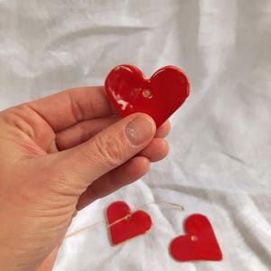 Könnte beinhalten: Eine Hand hält ein glänzendes, rotes Keramikherz mit einem kleinen Loch oben. Zwei weitere rote Herzen, ebenfalls mit Löchern, sind an einer dünnen, naturfarbenen Schnur aufgereiht. Die Herzen befinden sich auf weißem Hintergrund.