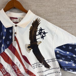 May include: A white polo shirt featuring a large bald eagle in flight, a formation of four fighter jets, and a graphic of the American flag. The shirt includes the text We the People and additional script text printed across the fabric.