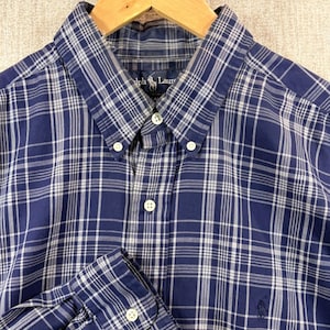 May include: A button-down shirt in a blue and white plaid pattern. The shirt has a button-down collar and a buttoned cuff. The shirt is made of a lightweight fabric and has a classic fit. The shirt is on a wooden hanger.